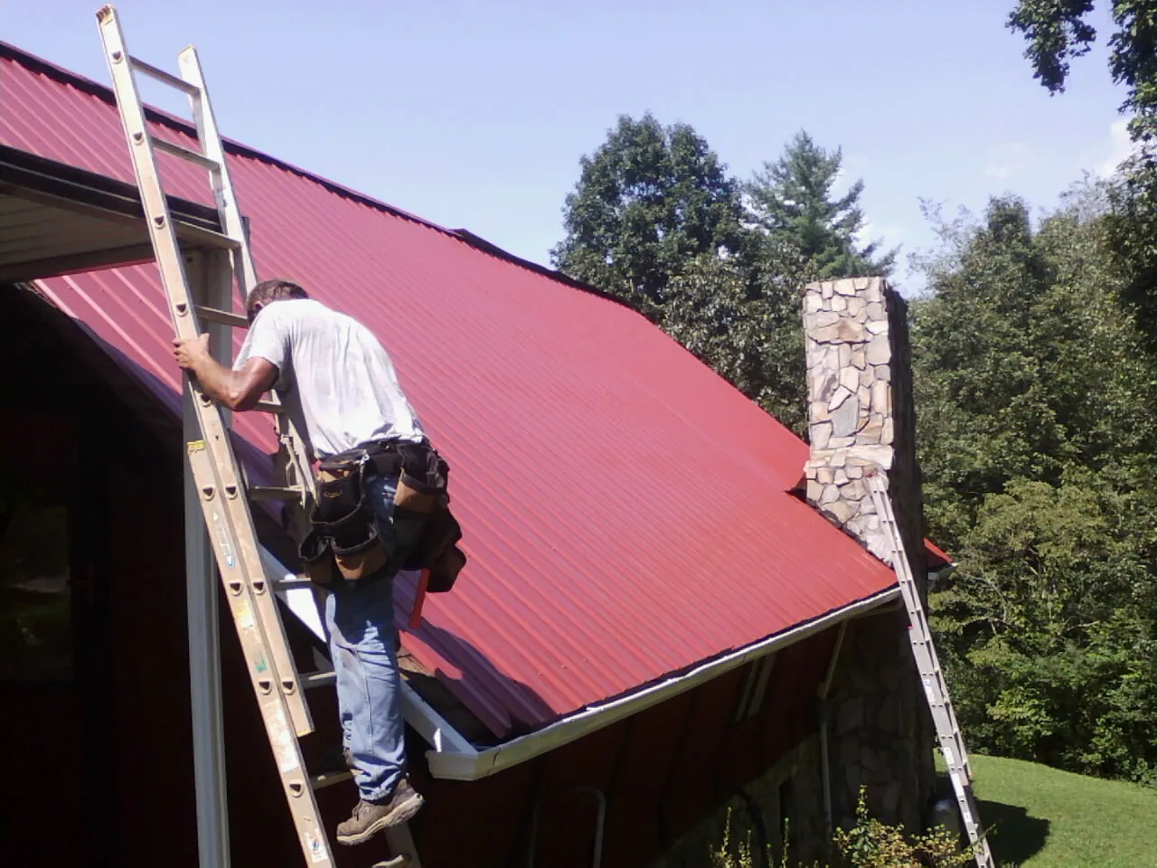 Roofer on a ladder beside a red metal roof and stone chimney — Soffit & Fascia Repair by Roofing Brothers in Eagleville